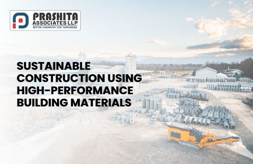 A wide-angle view of a modern construction material plant with stacks of concrete pipes, silos, and machinery. The foreground shows a bright orange industrial vehicle, while the background features a clear sky with light clouds and a distant mountain range. Overlaid text reads: "Sustainable Construction Using High-Performance Building Materials" with the Prashita Associates LLP logo in the top-left corner.